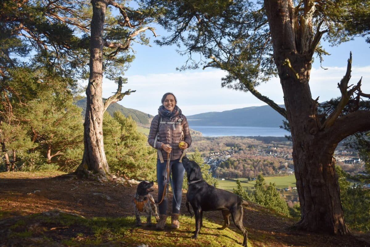 A woman stands on a wooded hilltop with two dogs - a black Labrador and a speckled Spaniel - overlooking a wide valley. In the background, a large loch stretches toward distant mountains under a clear blue sky.